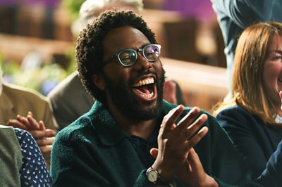 A character called Gus (Daniel Lawrence Taylor) laughs and claps.