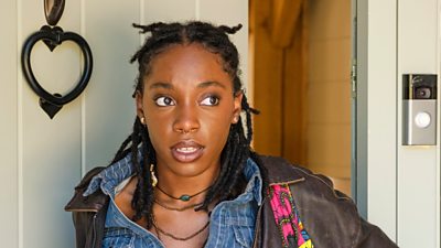 A woman (Leah, Boarders) in a backpack comes through a door. 