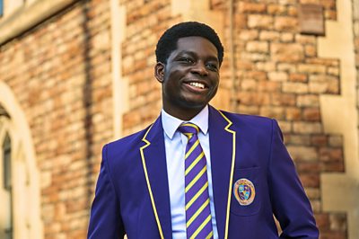 A young student (Femi in Boarders) in a purple uniform smiles outside a wall 