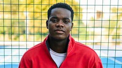 A man in a red tracksuit top (Jaheim from Boarders) poses outside a gate.