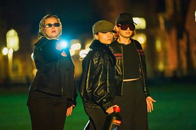 Three young people dressed in black look on with dark glasses and torches. L-R: Beatrix (Tallulah Greive), Florence (Rosie Graham) and Yelena (Ruxandra Porojnicu).