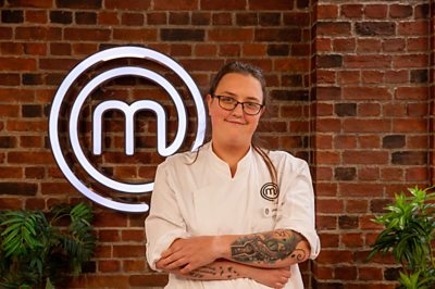 A woman with tattoos and chef whites poses in the MasterChef studios