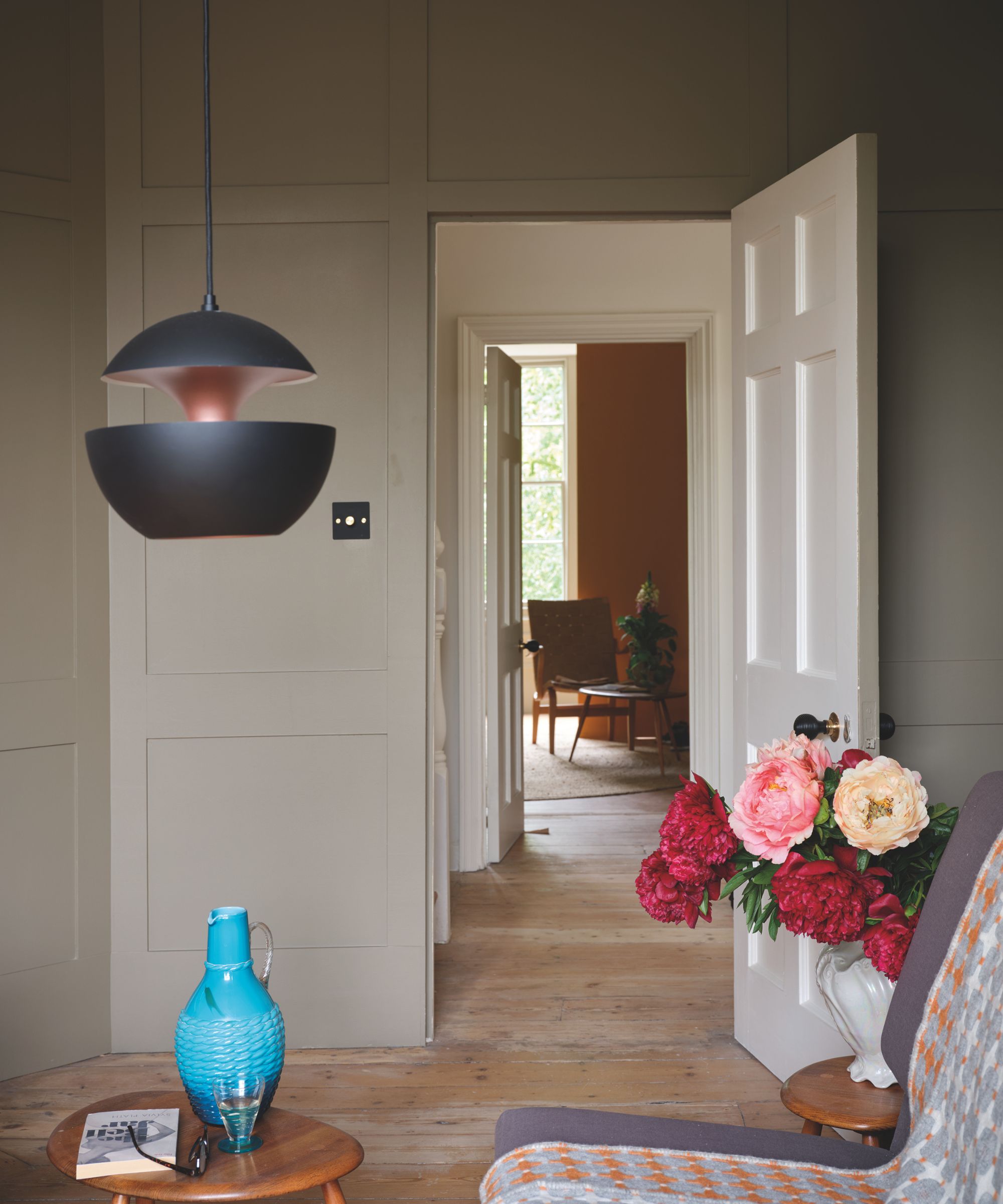 Living room with panelled walls painted in taupe brown, daybed with a wool throw on top, Ercol side tables with a blue vase and another with a vase of peonies, with a midcentury black low pendant above