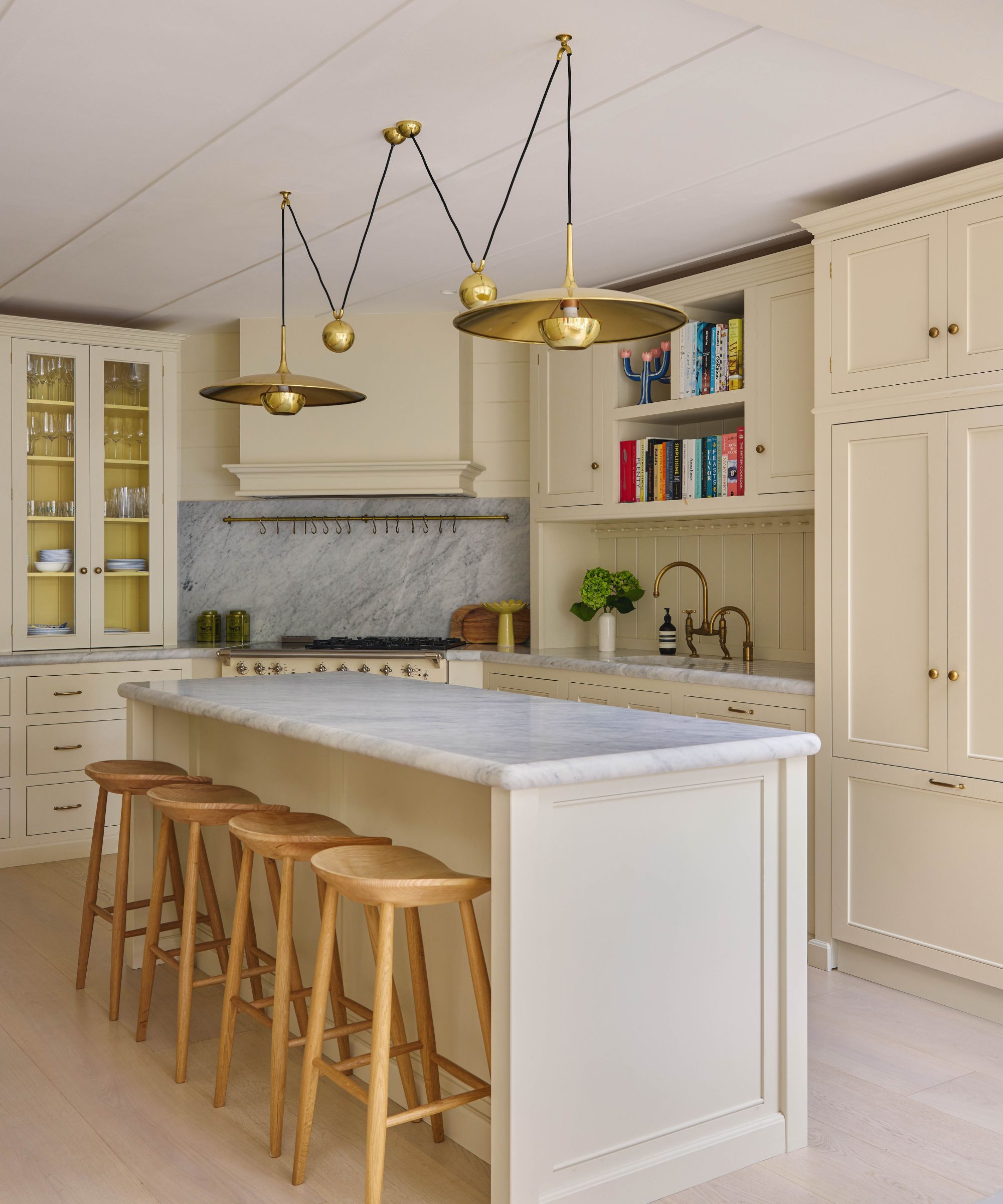 A sophisticated cream kitchen with a marble-topped island and four wooden barstools. Unique brass "pulley-style" pendant lights hang above the island, set against a marble backsplash and open shelving filled with colorful books.