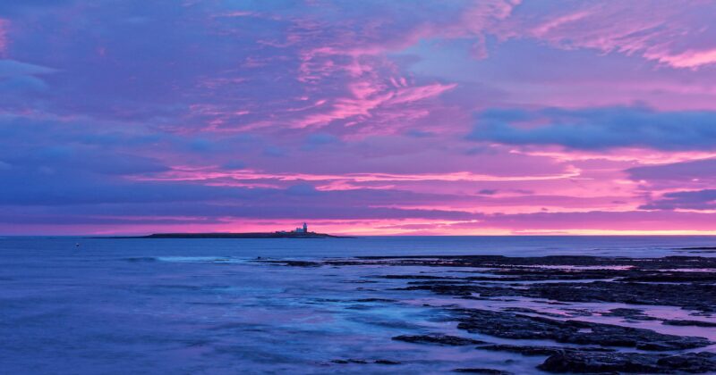 A rocky shoreline at dusk with dramatic pink and purple clouds in the sky. A small island with a lighthouse sits on the horizon, surrounded by calm ocean water reflecting the colorful sunset.