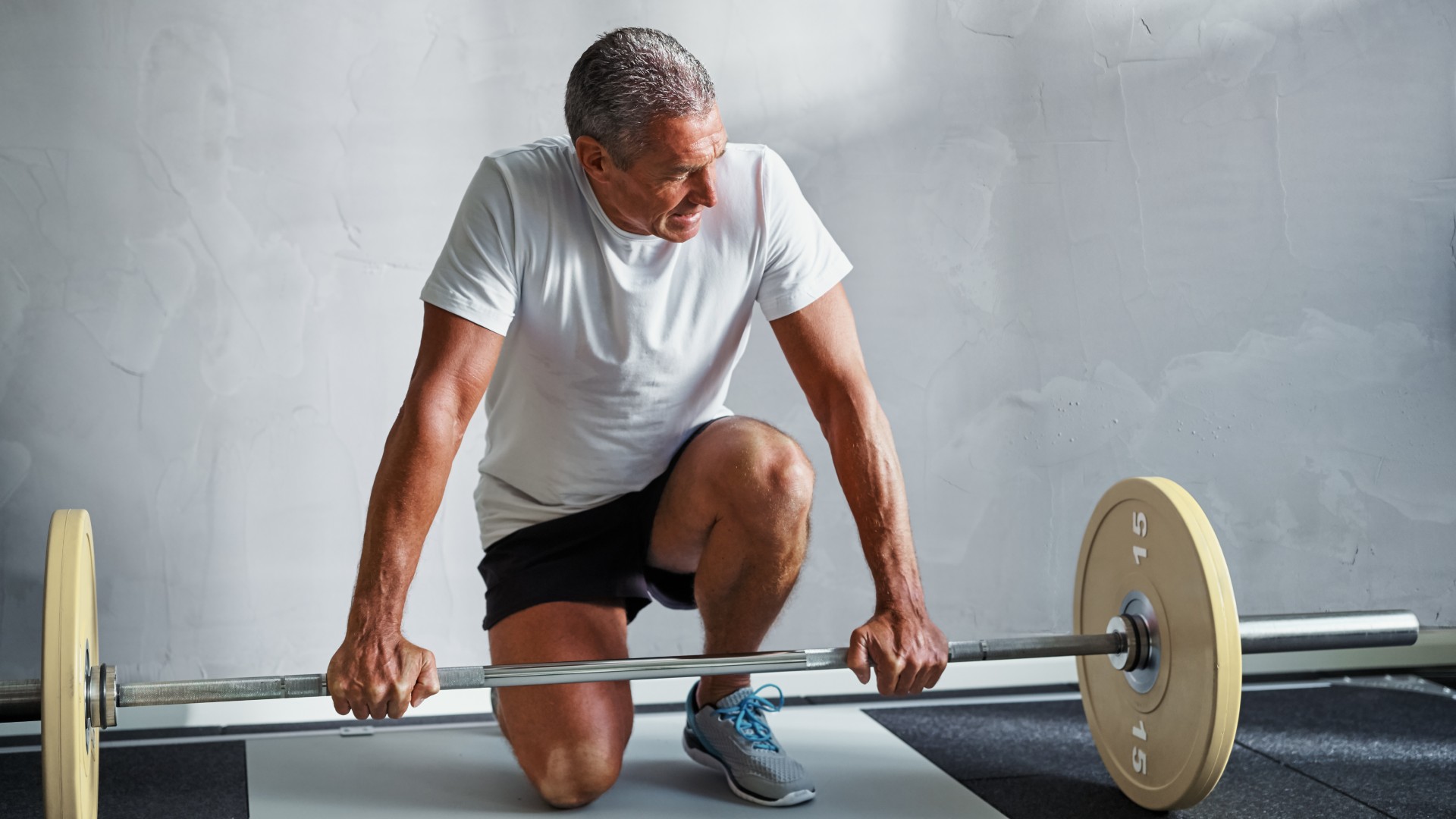 Older man in activewear crouching next to a barbell in gym