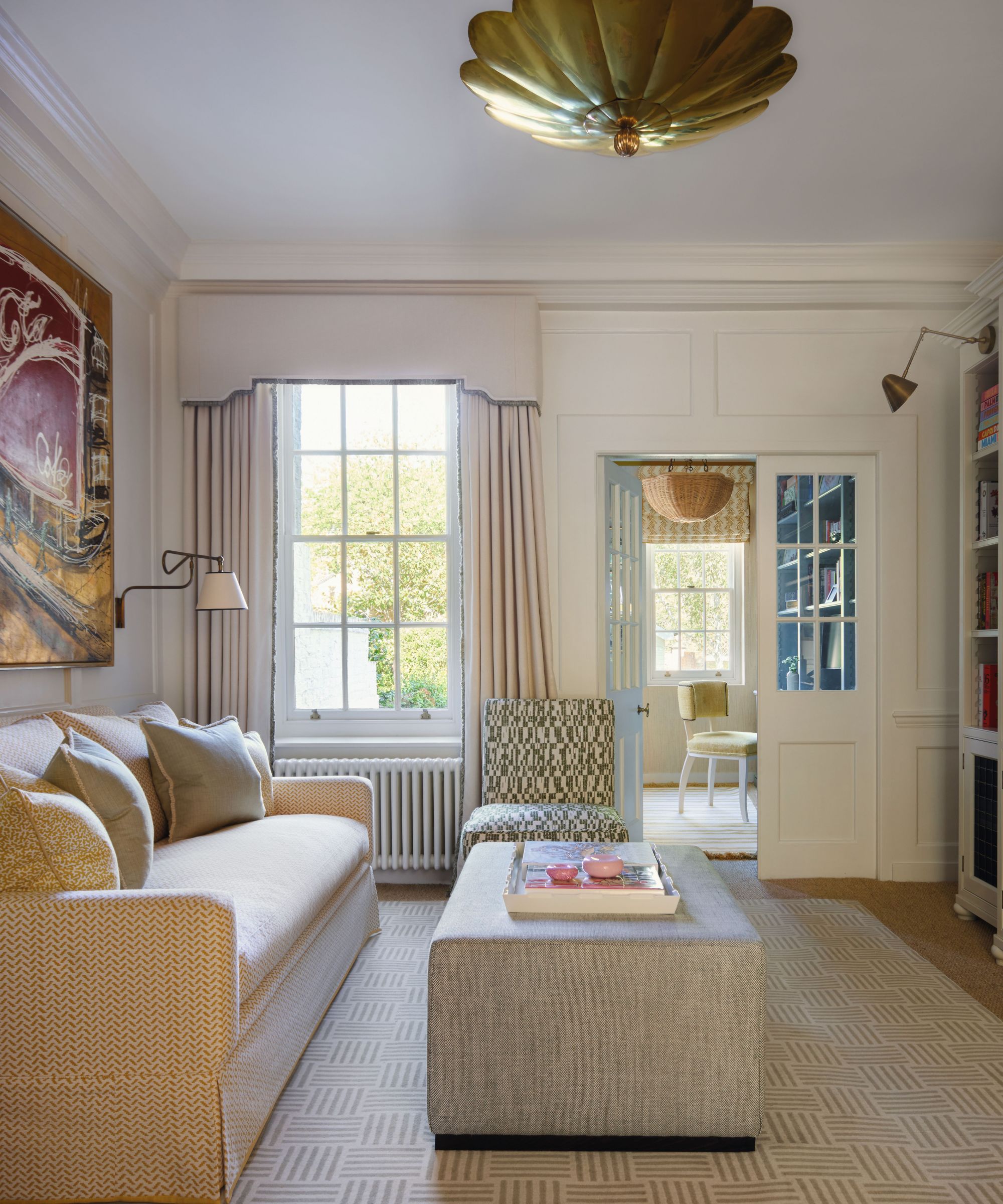 A cozy sitting room with a cream patterned sofa, a large grey ottoman, and a statement gold scalloped ceiling light. A doorway leads to a sunlit home office with a yellow chair.