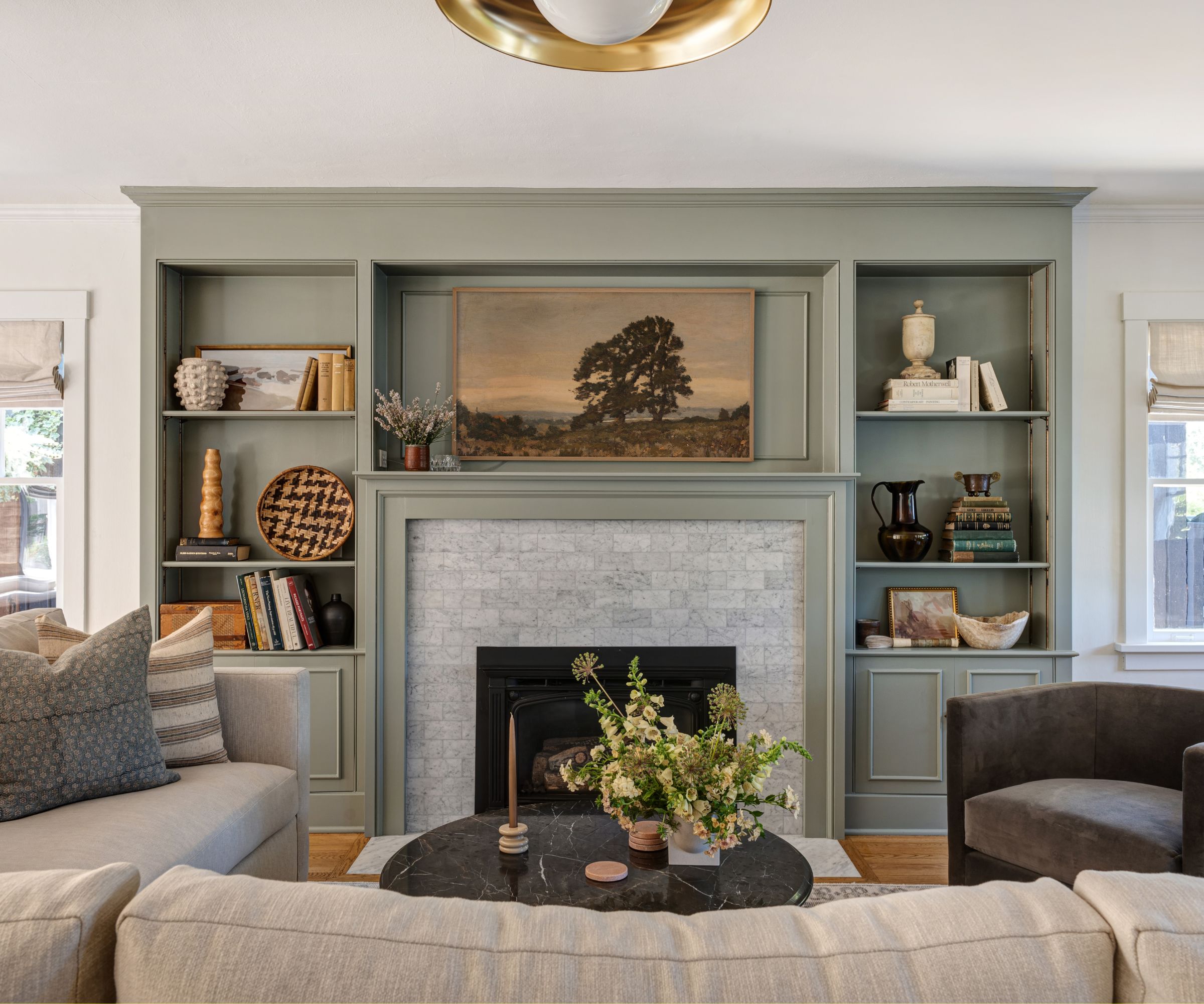 Living room with grey-green painted built-in bookshelves with decor and books, fireplace with a landscape painting above it, neutral sofa, and a dark grey velvet armchair