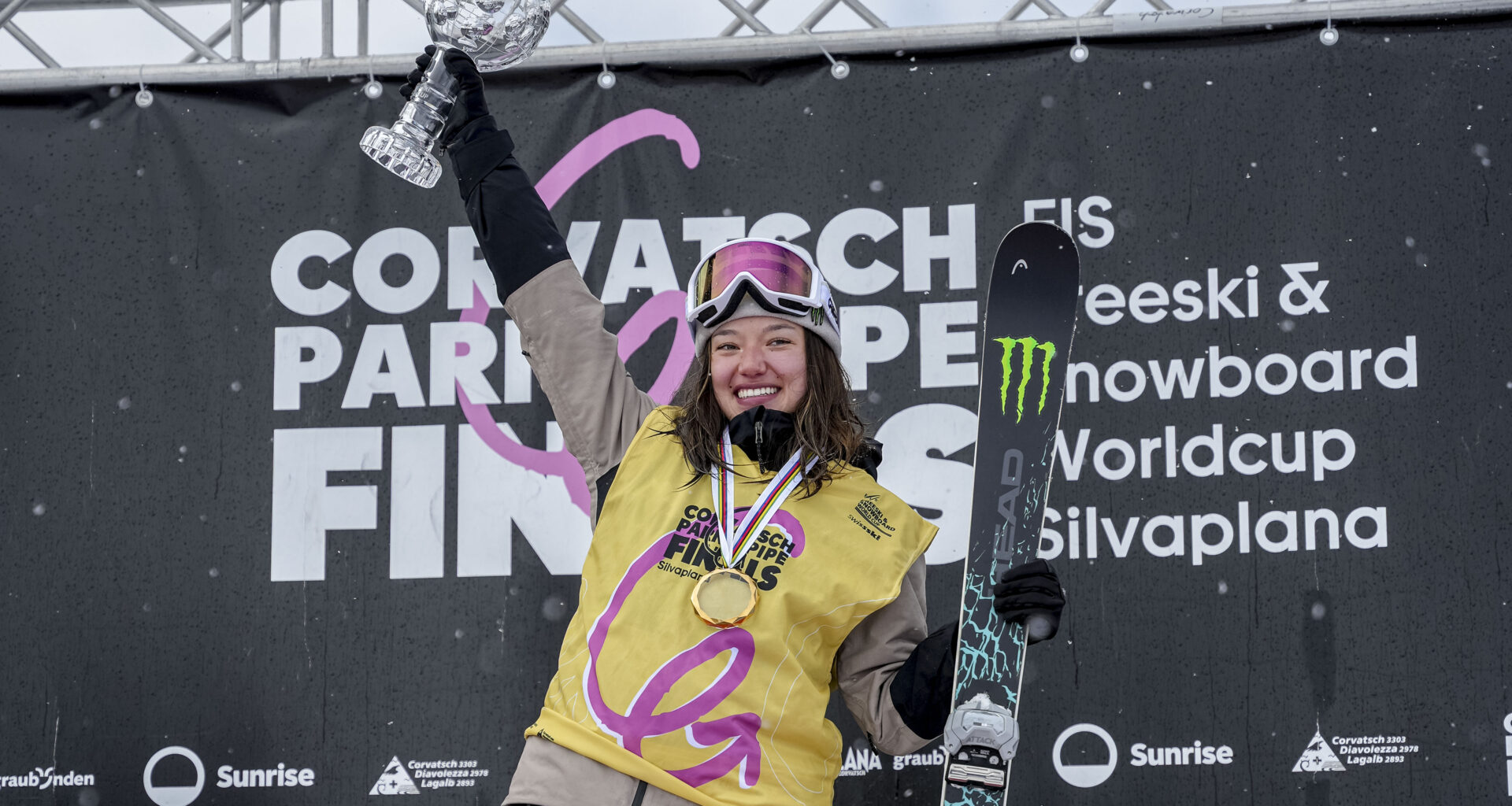 Two skiers smiling and holding crystal trophies, dressed in winter gear with medals around their necks, standing on a snowy slope.