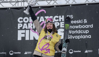 Two skiers smiling and holding crystal trophies, dressed in winter gear with medals around their necks, standing on a snowy slope.