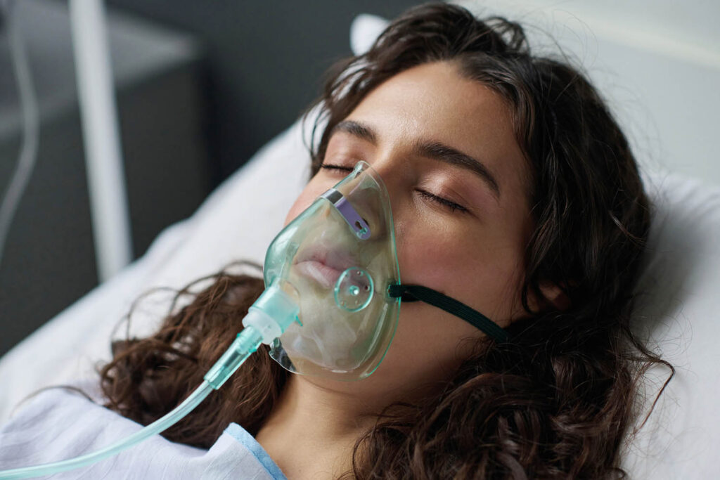 Close-up of young woman lying on bed with her eyes closed and breathing with oxygen mask in hospital