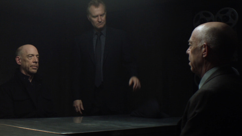 Howard Silk sits across an interrogation table facing his double, Howard Silk, while Josef Aldrich oversees the interrogation in 