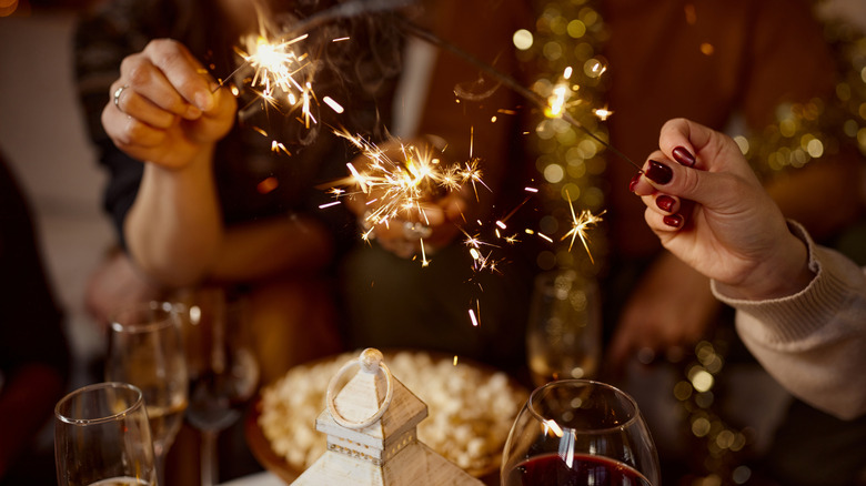 Hands hold small fireworks over wine glasses and table