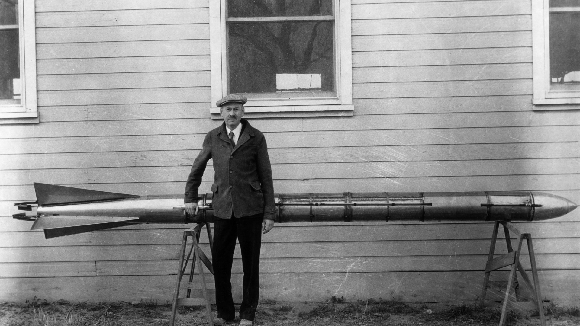 a man in a suit poses in front of a long, slender metal tube beside a wood-paneled building
