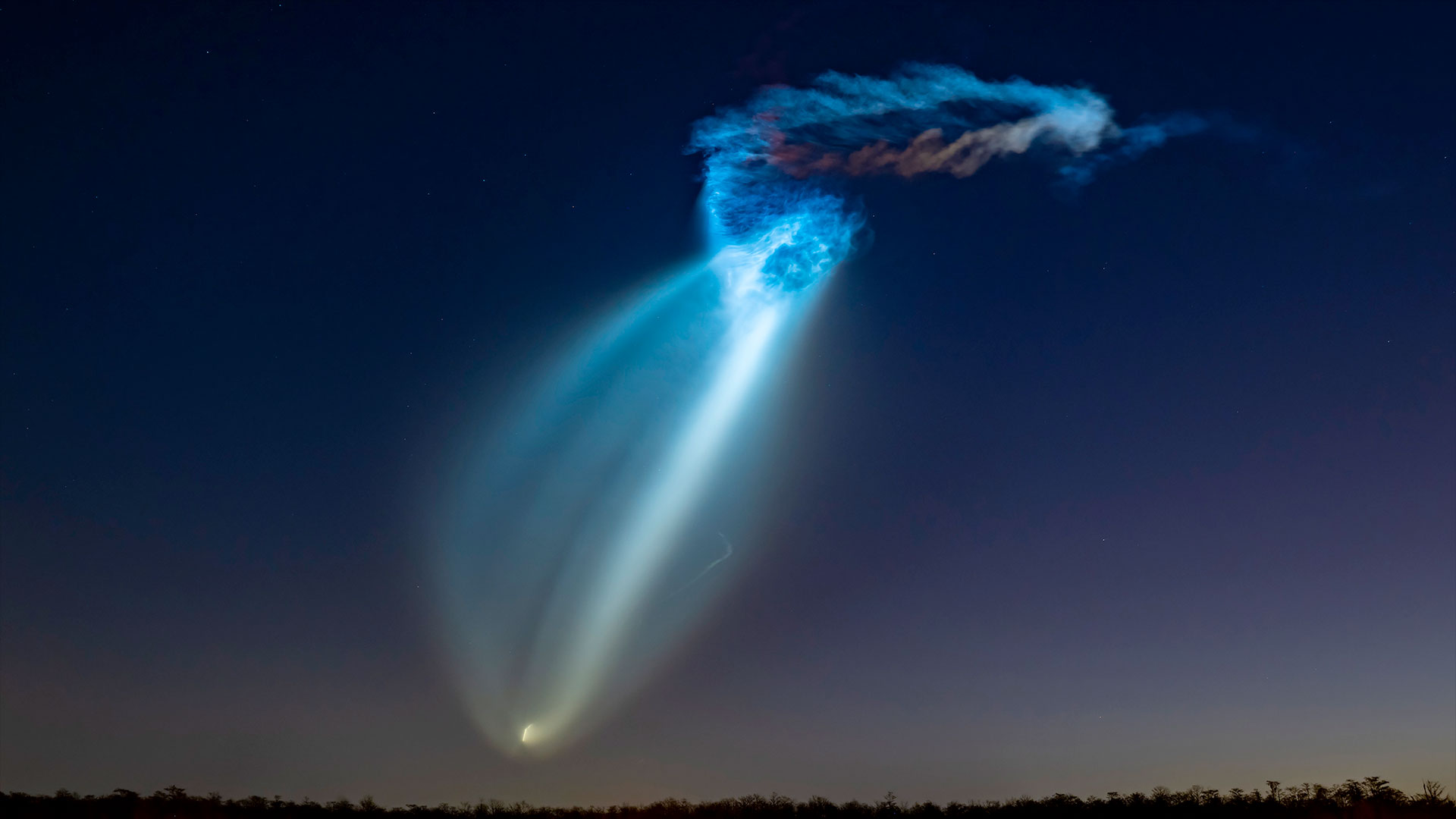 a rocket's high-altitude plume is lit up in shades of blue and orange by the sun prior to sunrise.