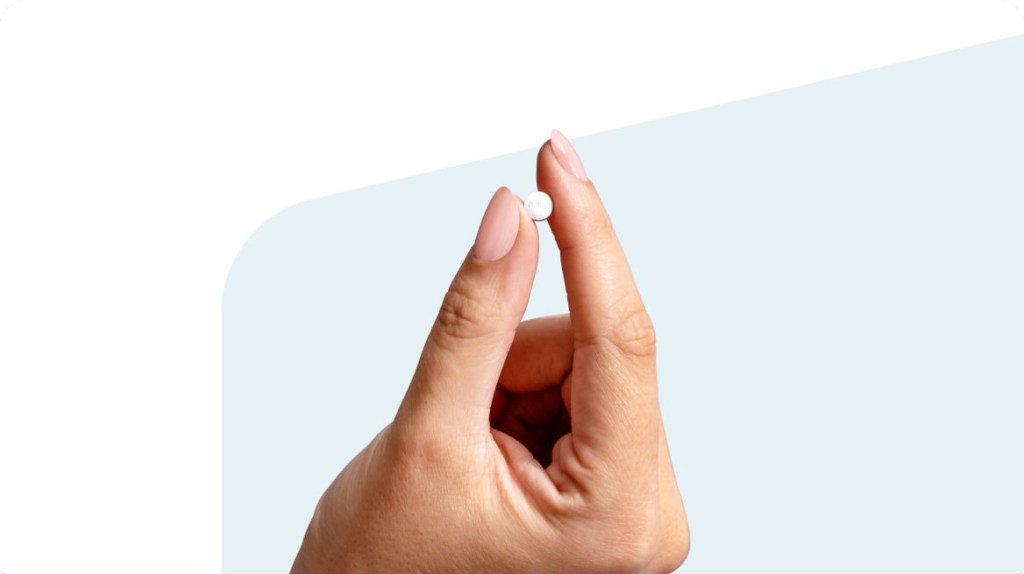 A hand holds a small white pill between its index finger and thumb.
