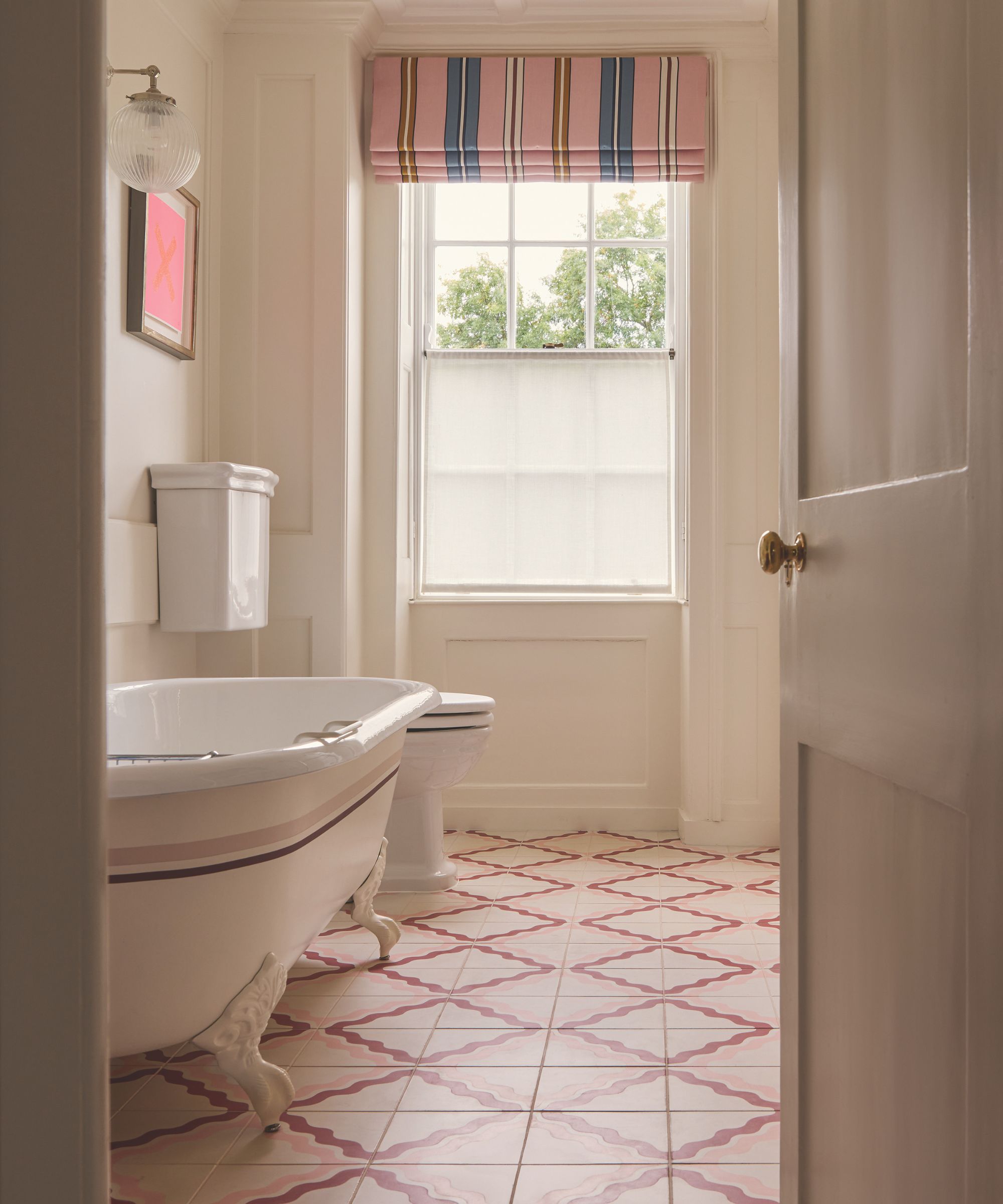 A charming traditional bathroom seen through an open doorway. It features a white clawfoot bathtub with a pink stripe, pink-and-white patterned floor tiles, and a window dressed with a pink, blue, and gold striped Roman blind.