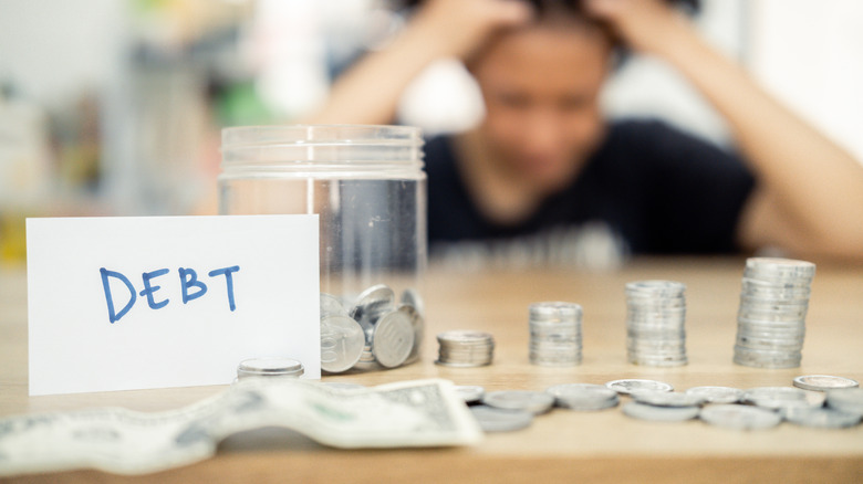A woman sits at a table, looking distressed, with piled coins and a debt note.