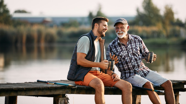 Happy senior fisherman and his adult son talking while having a drink on a pier.