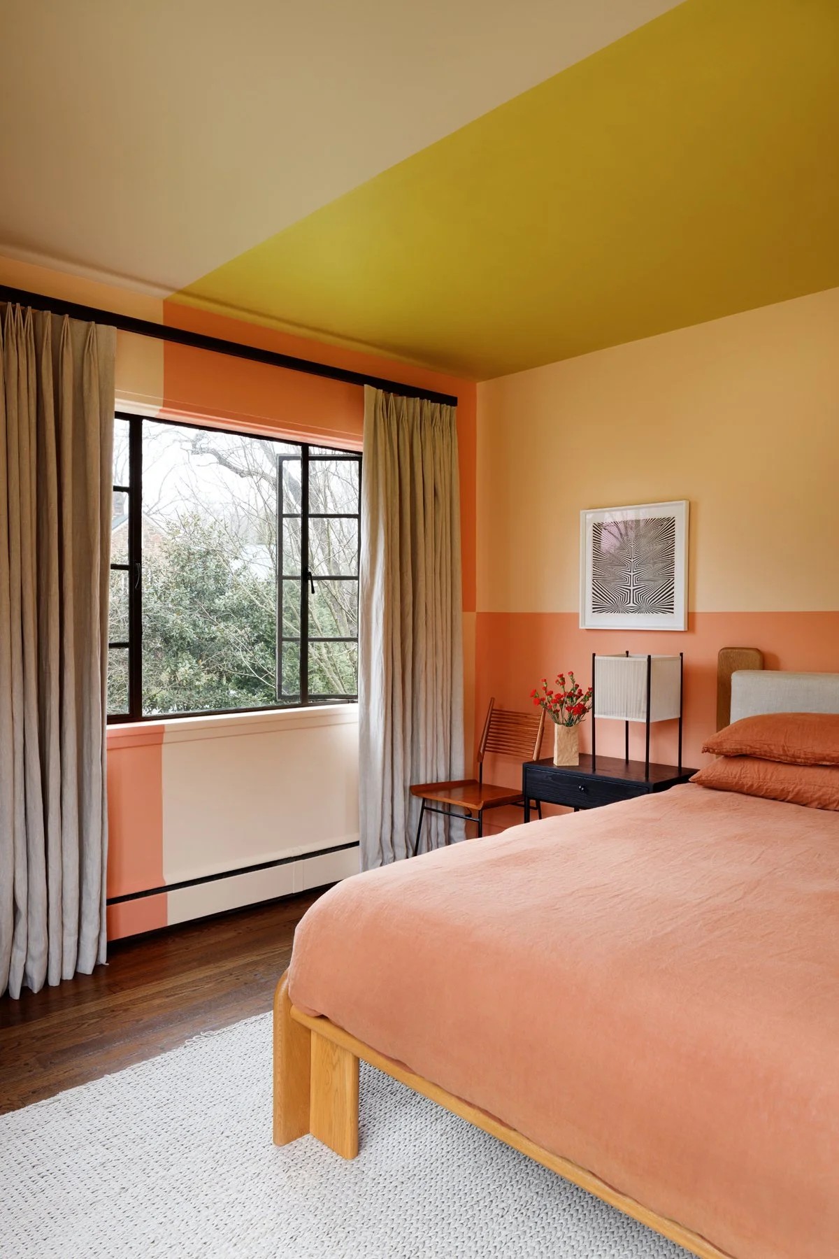 Image of a bedroom in a peach color block with a lime green and white color block ceiling. The bed has peach bedding and there is a black side table with a table lamp on it.