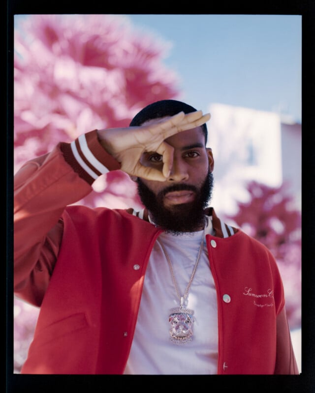 A bearded man in a red jacket and white shirt stands outdoors, holding his hand up to his face making a circle with his fingers. He wears a large pendant necklace. The background features pink foliage.
