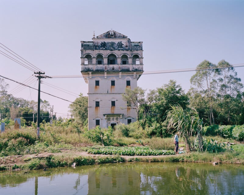 A tall, multi-story building with arched windows and decorative details stands near a pond, surrounded by greenery and vegetable plots, with a person walking along the water's edge on a sunny day.