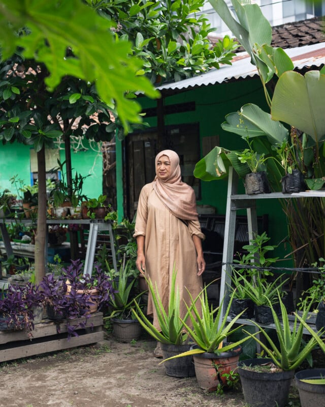 A woman in a beige dress and hijab stands in a lush garden, surrounded by potted plants and greenery, with a green building and trees in the background.