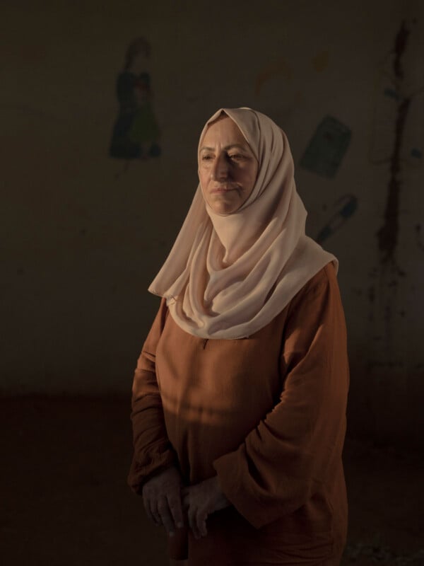 A woman wearing a light-colored hijab and an earthy brown dress stands in soft light against a dim, plain wall with faint, blurred drawings in the background. She gazes slightly downward with a thoughtful expression.