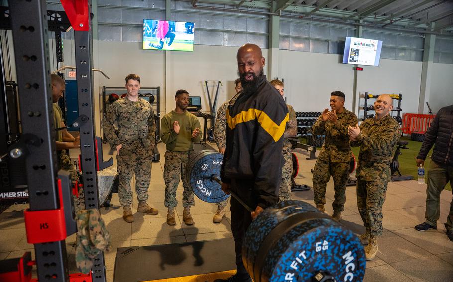 A man lifts a loaded barbell as Marines cheer during a strength training demonstration inside the gym.