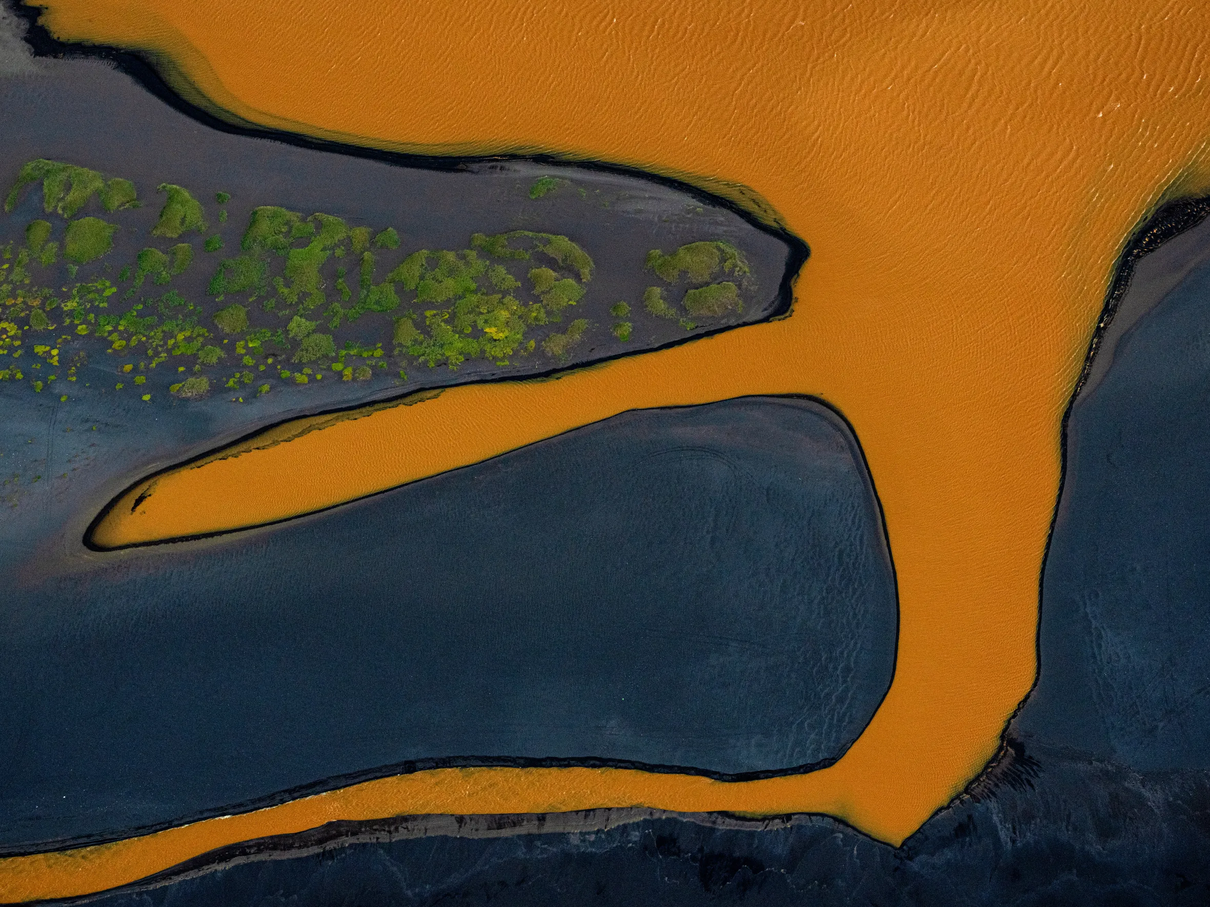 An aerial view of a winding orange river flowing through dark land with patches of green moss.
