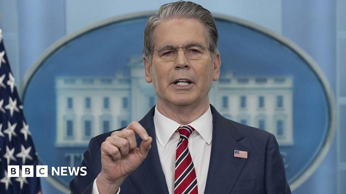 Face and shoulders of US Treasury Secretary Scott Bessent wearing a blue suit, white shirt, and red, white, and navy blue striped tie with a US flag lapel pin addressing the press, who are not in shot, in the White House press room. Behind him, there is US flag and and, mostly blocked by his face and body, a big image of the White House with the text 'The White House' beneath.