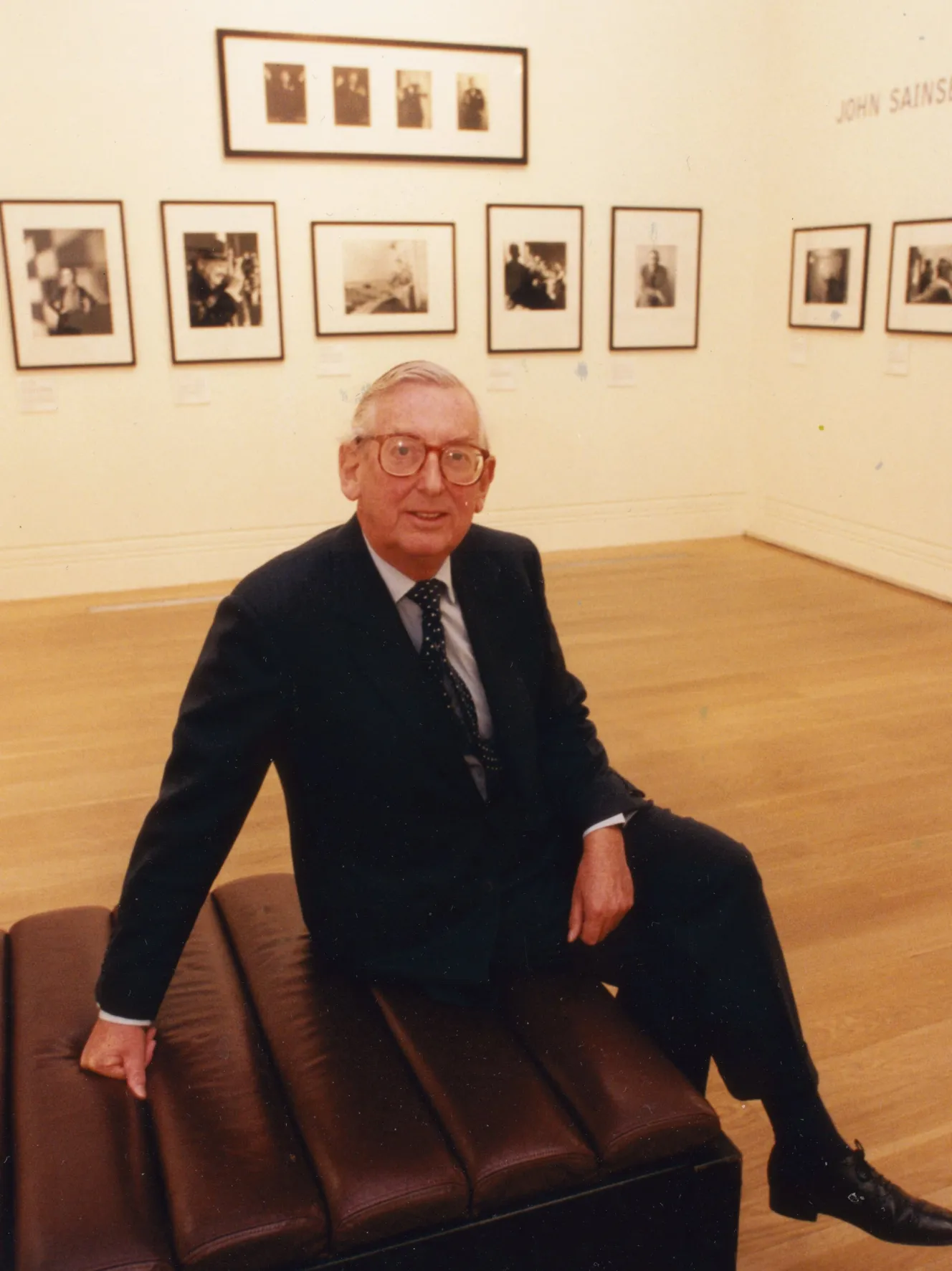 Lord Sainsbury sitting on a bench in front of an exhibition of photographs.