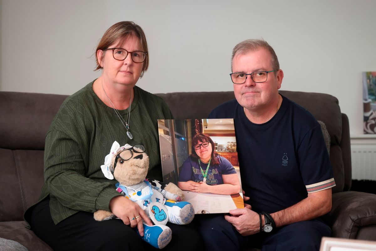 Mid-aged woman with short hair and glasses (left) in green jumper and man with short hair and glasses in dark navy t-shirt sat on sofa holding picture of young woman with glasses at cafe/restaurant table