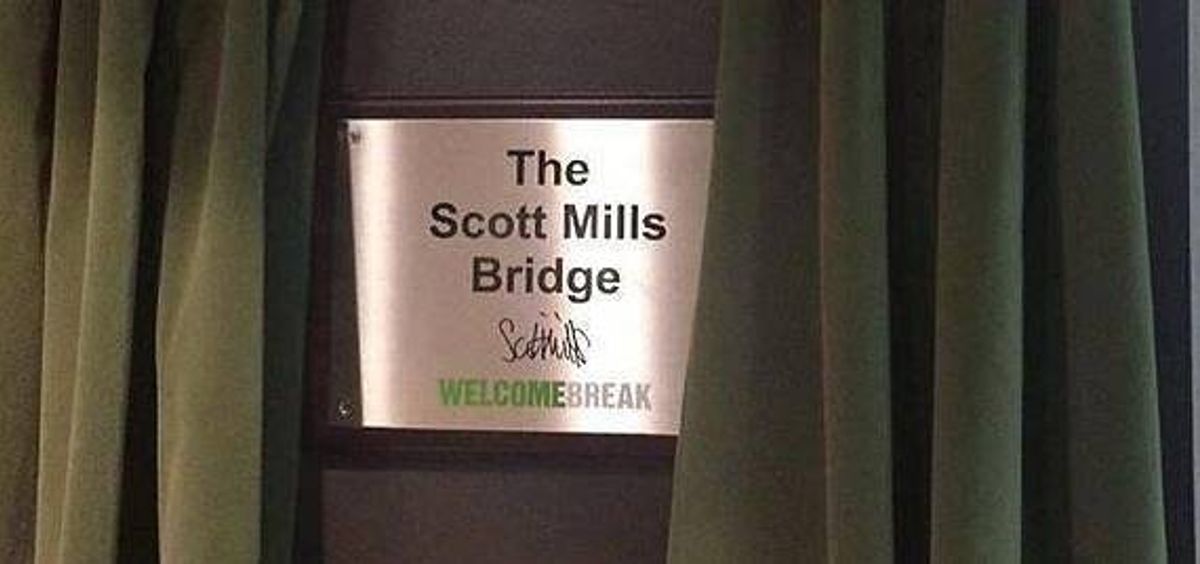 An image displays a metallic plaque with the inscription "The Scott Mills Bridge," positioned between two dark green curtains, suggesting an official or commemorative marker.