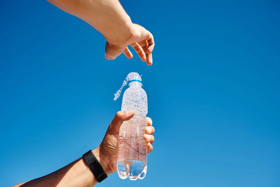 Two hands open a sparkling water bottle under a clear sky. Bubbles are visible, capturing a refreshing moment