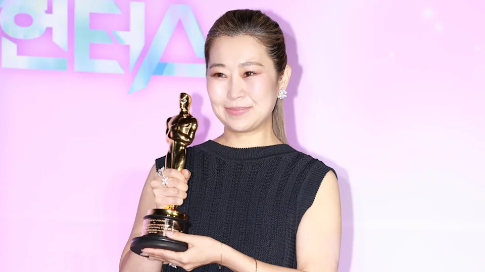'KPop Demon Hunters' co-director Maggie Kang in a black dress holds a gold trophy, smiling against a pink background.