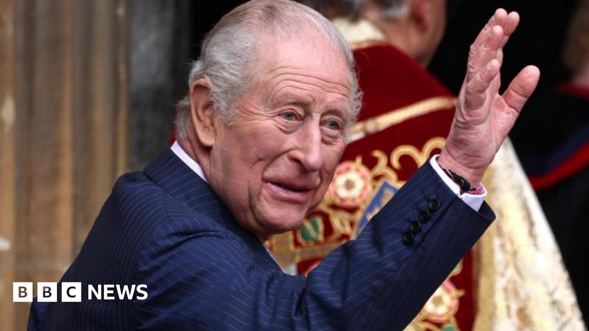 King Charles, dressed in a blue pinstriped suit, is smiling and waving at the entrance of St. George's Chapel, Windsor