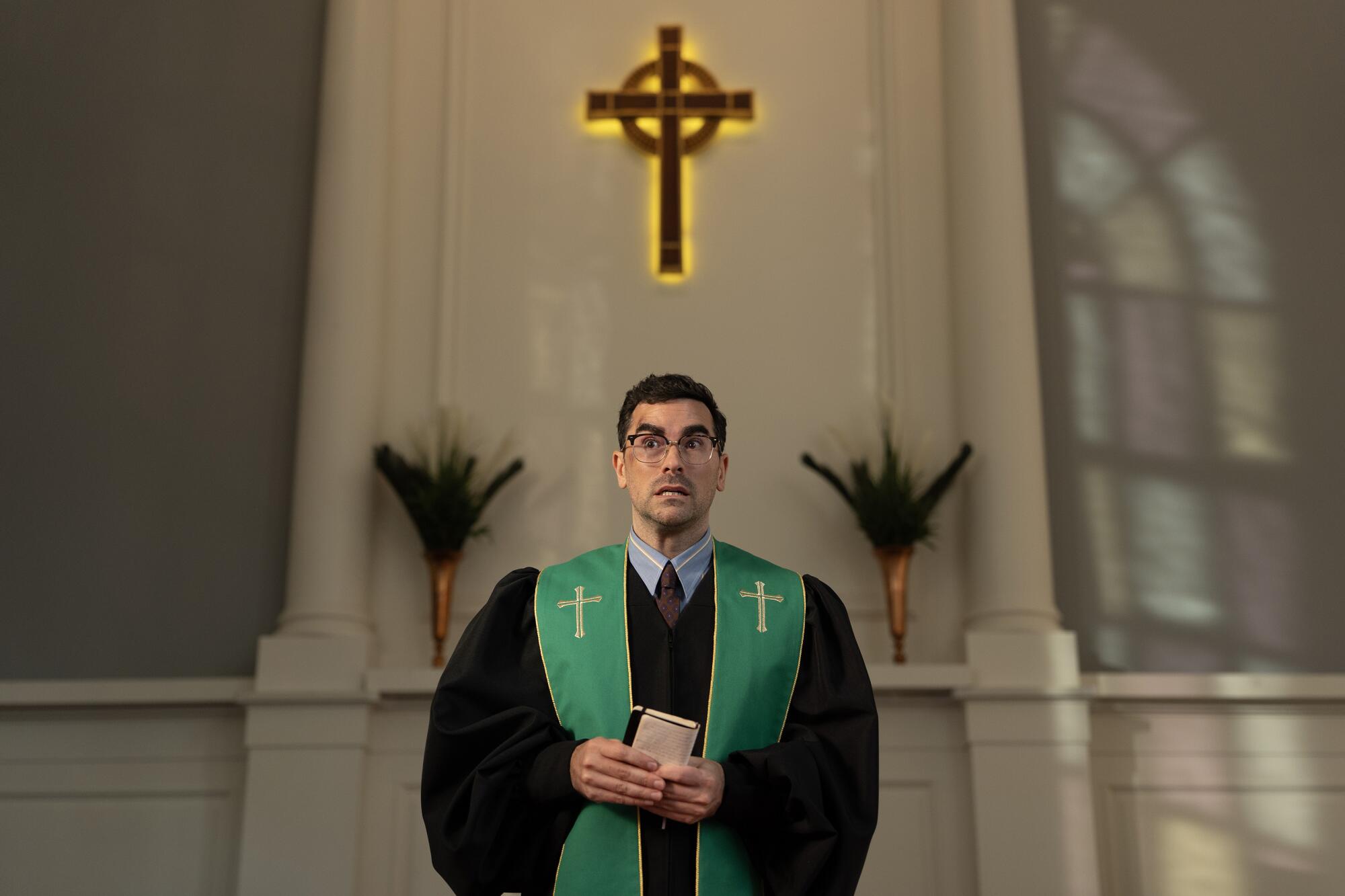 A man in a black cassock and green stole holds a notebook in his hands while standing on a pulpit. A cross hangs behind him.