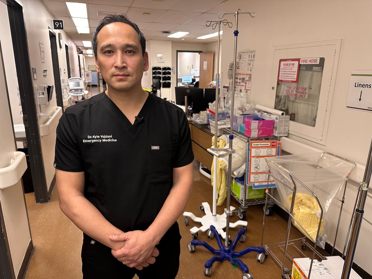 A middle-aged doctor in black scrubs holds his hands in front of him and looks at the camera with a neutral expression in an empty hospital corridor, with supplies along the wall.