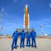 Four Artemis II crew members, dressed in blue jumpsuits, pose in front of a rocket.