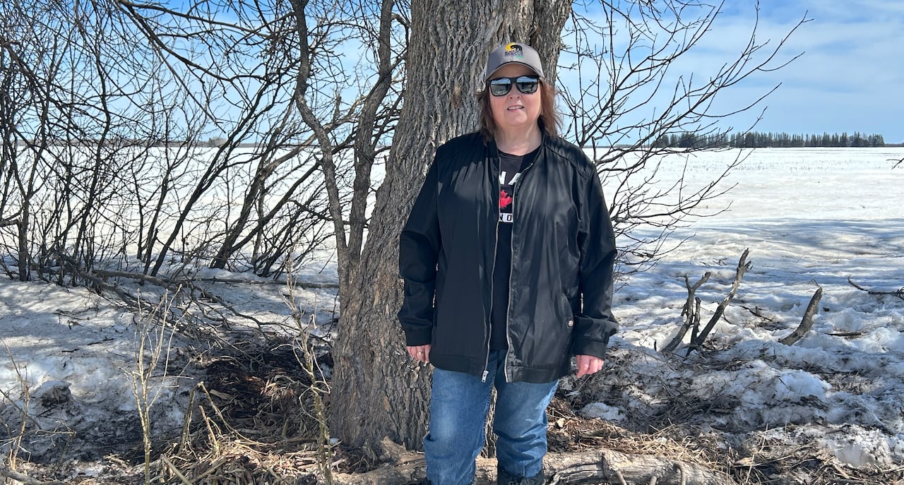 Woman wearing black spring jacket, blue jeans, a baseball cap and sunglasses while standing infront of a tree with snow on the ground