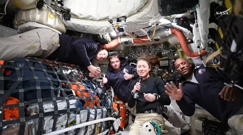 Four Artemis II astronauts in a spacecraft cabin, surrounded by equipment, smile and give thumbs up, with one holding a microphone—radiating hope as they prepare for their historic journey to the moon and look forward to seeing Earth from afar.