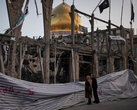 People look at a destroyed building within the Grand Hosseiniyeh, with the mosque visible in the background, after it was hit in airstrikes. 