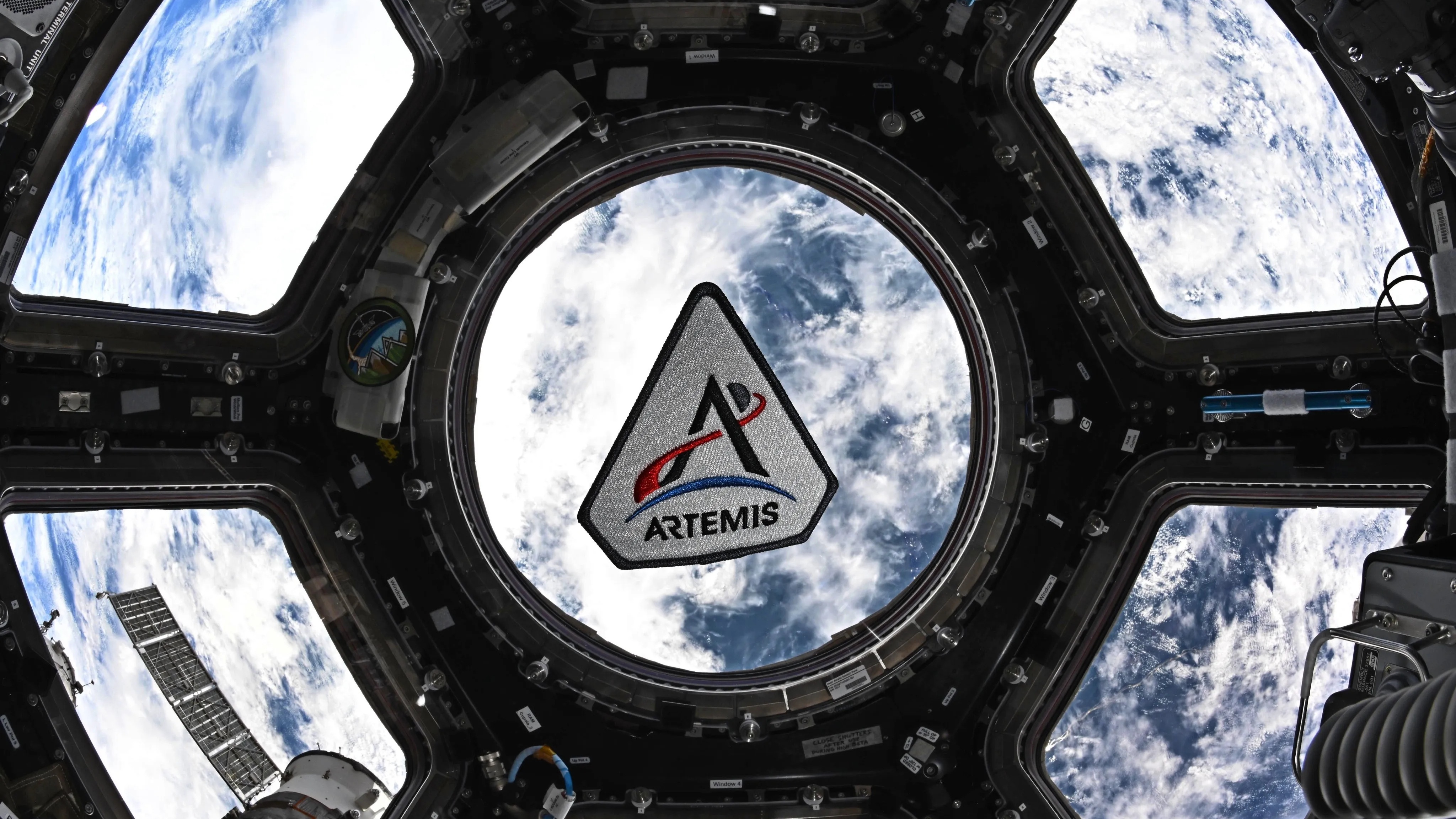The ISS cupola window with an artemis patch in the center section of the window. Earth is seen behind.