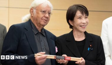Japanese Prime Minister Sanae Takaichi poses with Ian Paice. They hold a set of signed drumsticks between them. Takaichi has short black hair and wears a black blazer over a black top. Paice has long white hair, tied back, and wears glasses, a black blazer and a grey buttoned shirt.