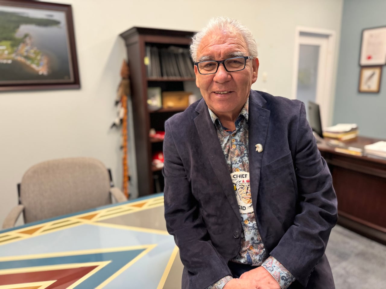Man in dress shirt, blazer and glasses leans against a table in an office