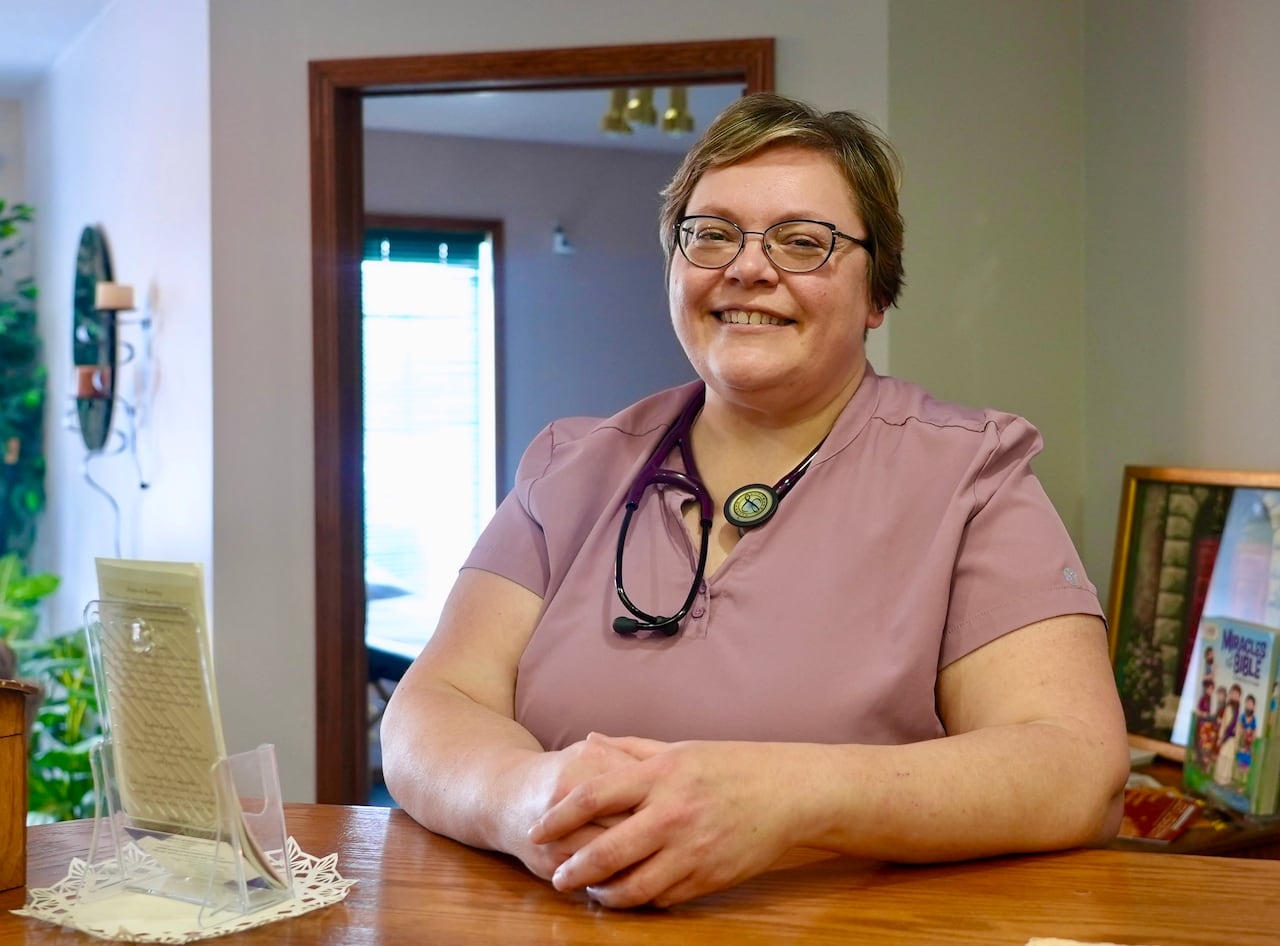 A lady wearing a stethoscope around her neck is smiling at the camera while leaning on a wooden counter.