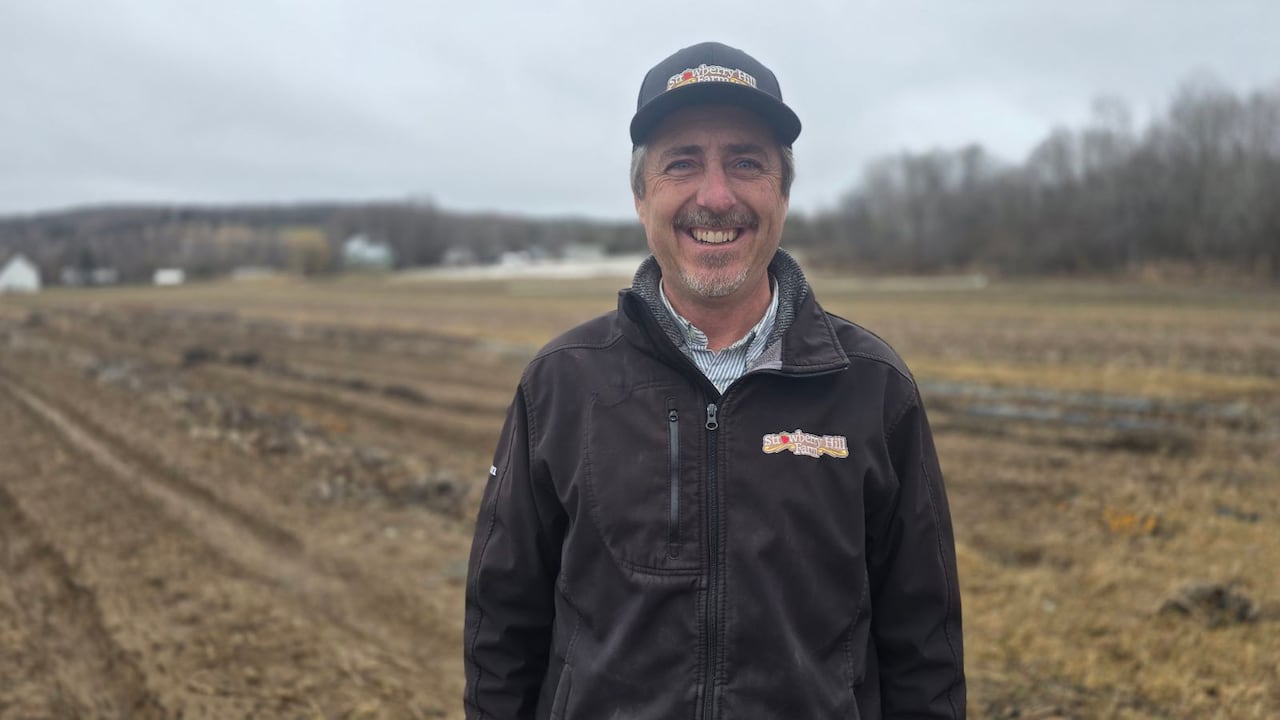 A man stands in a farm field, smiling on a cloudy day