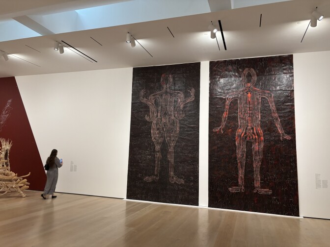 Two approx 16-foot-tall paintings on a white museum gallery wall. Each are fully painted a shiny brown/black with a human-like figure formed by hand on each of them. The figure on the left is light brown and the one on the right is more red.