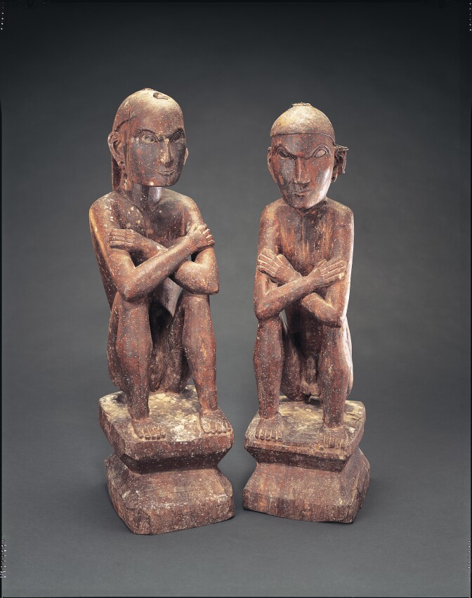 Two carved wooden figures of people sitting with their arms crossed and resting on their knees. They appear to have serious expressions on their faces and are unclothed.